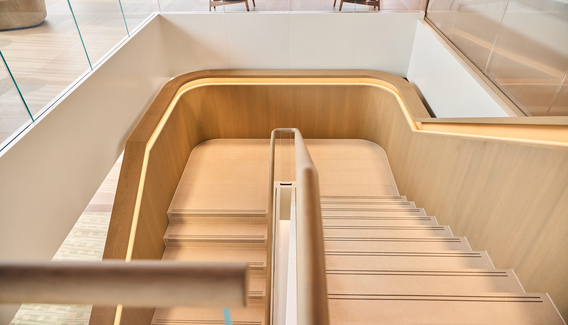 Modern office staircase with light wood-paneled walls, integrated lighting, glass railings, and a central handrail.