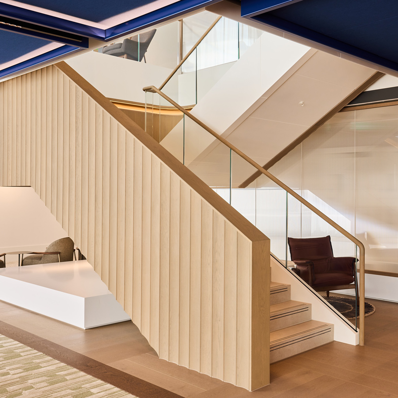 Modern office staircase with light wood paneling, glass railings, integrated lighting, and seating areas beneath a dark blue ceiling.