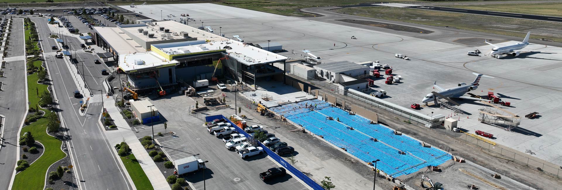 Aerial view of an airport terminal expansion under construction beside an active apron with parked aircraft.