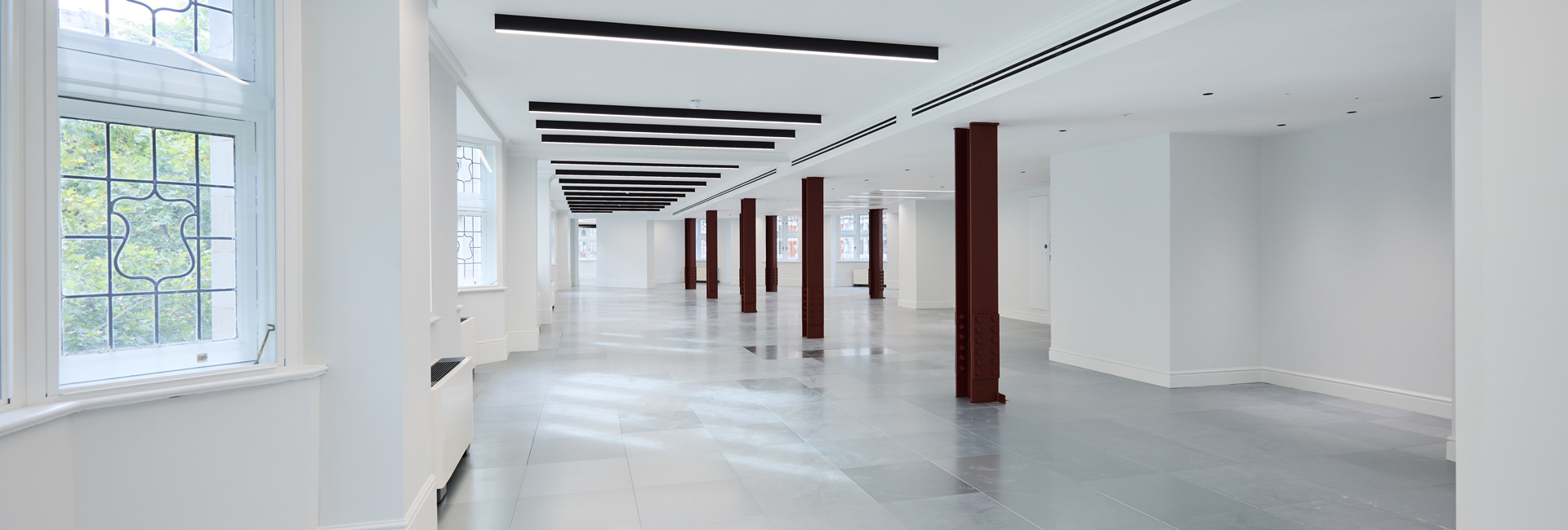 Spacious open-plan interior with decorative windows, gray flooring, linear ceiling lights, and exposed red columns.