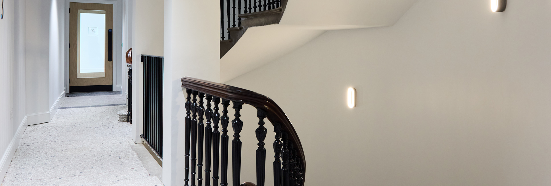 Renovated stairwell and corridor featuring ornate black balusters, a dark wood handrail, terrazzo flooring, and modern wall lights.