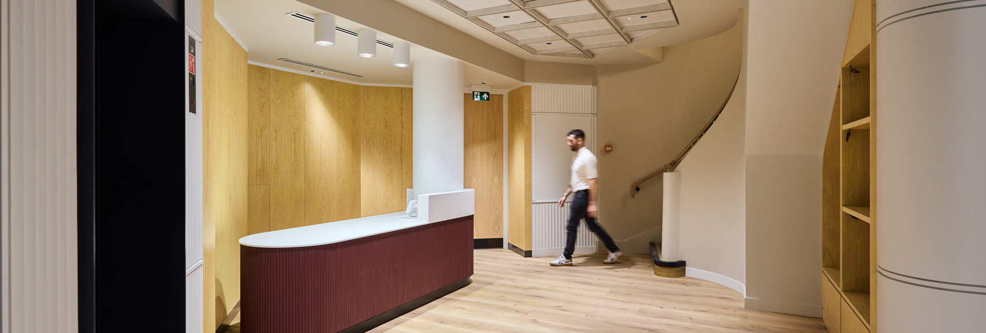Modern reception area with light wood finishes, a curved burgundy desk, and a person walking near the staircase.