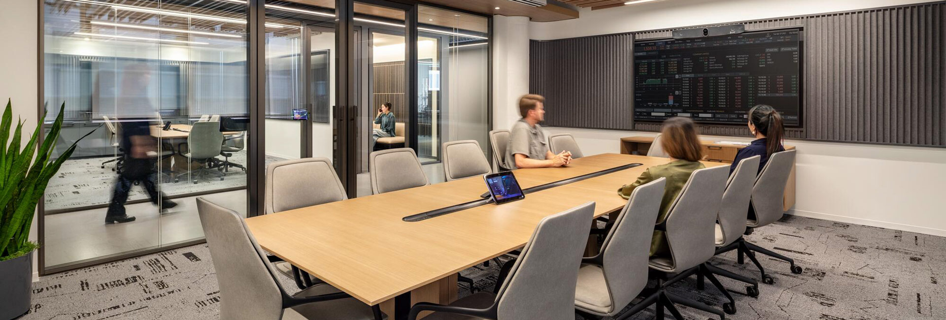 Employees meet in a glass-enclosed conference room featuring a long wood table and wall-mounted financial market display.