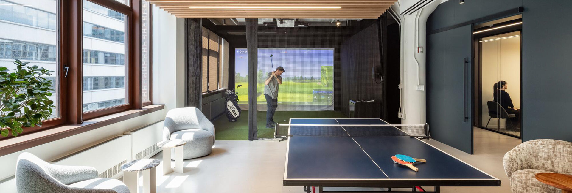 Sunlit employee game room featuring a golf simulator, table tennis table, and lounge seating.
