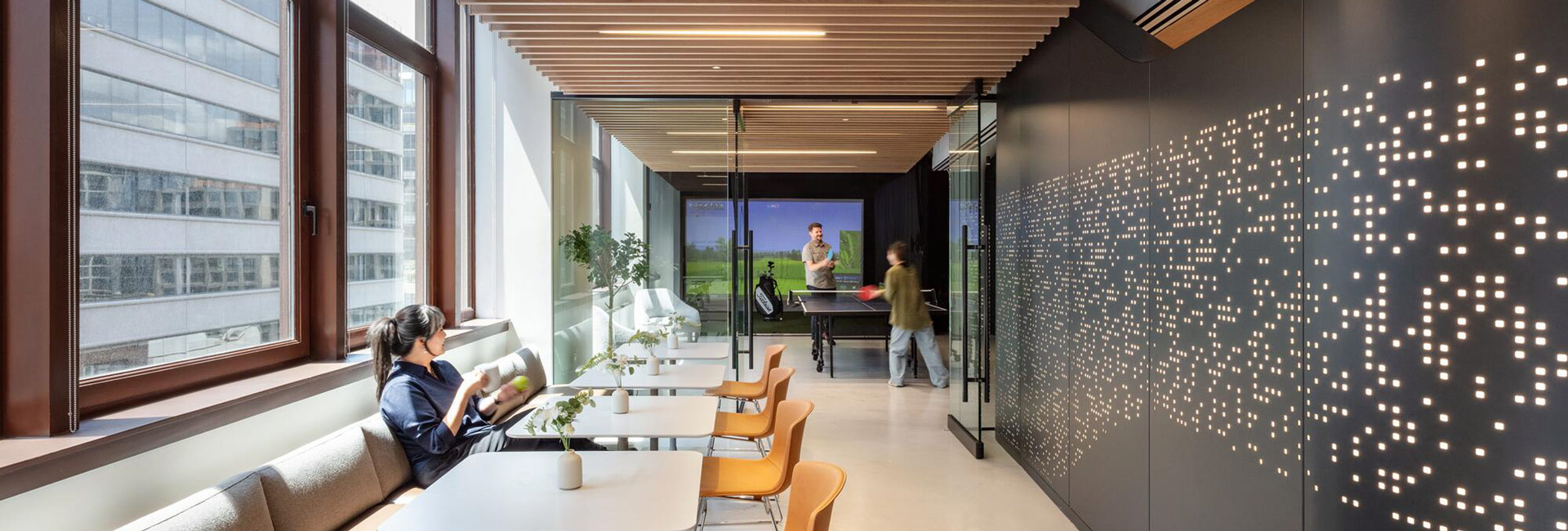Employee lounge with window-side seating, table tennis, a golf simulator, and an illuminated perforated feature wall.