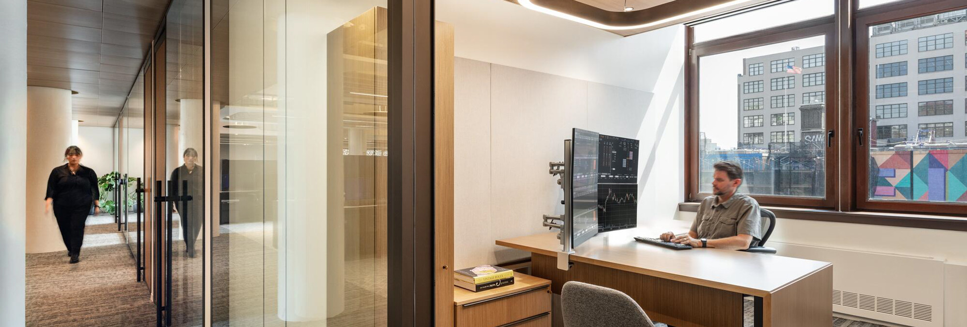 Private office with dual monitors displaying financial data, glass walls, wood furnishings, and large windows.