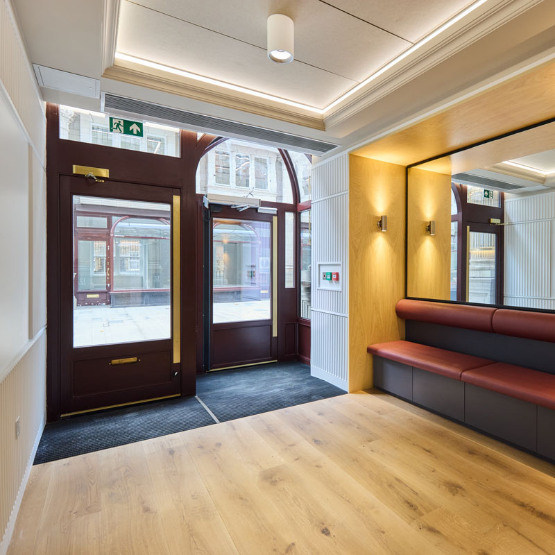 Modern building entrance with burgundy-framed glass doors, wood paneling, wall sconces, and a built-in red leather bench beneath a large mirror.