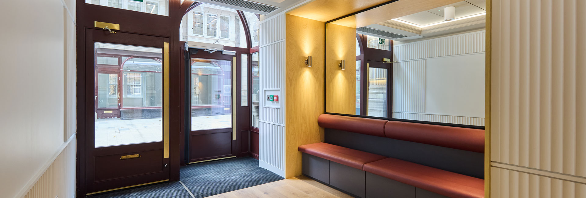 Modern building entrance with burgundy-framed glass doors, wood paneling, wall sconces, and a built-in red leather bench beneath a large mirror.