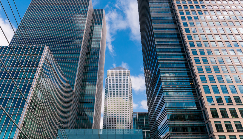 Modern glass-and-steel office towers rise against a bright blue sky in a dense urban business district.