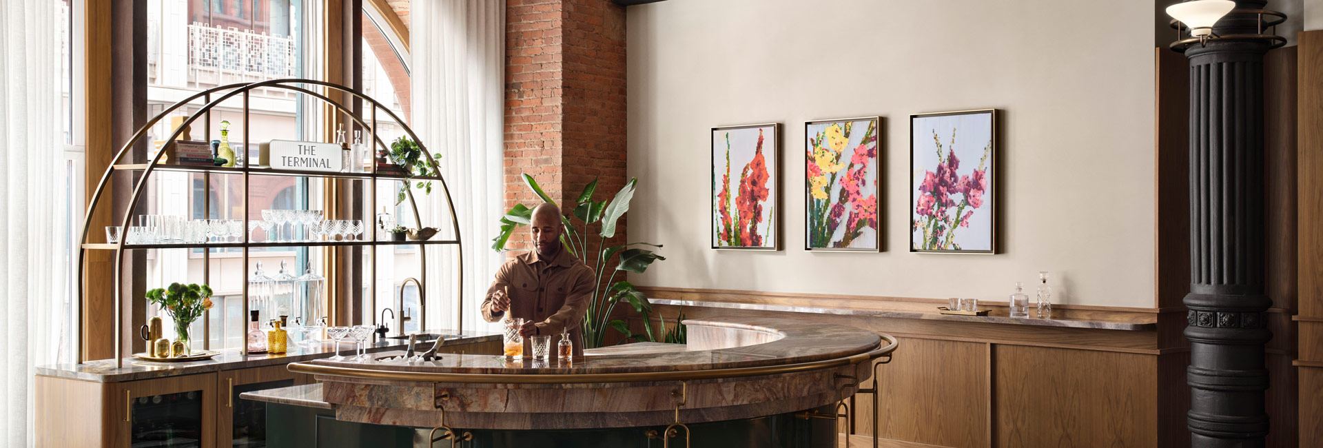 Open workspace with individual workstations, exposed brick, black columns, arched windows and abundant plants, with two people visible.