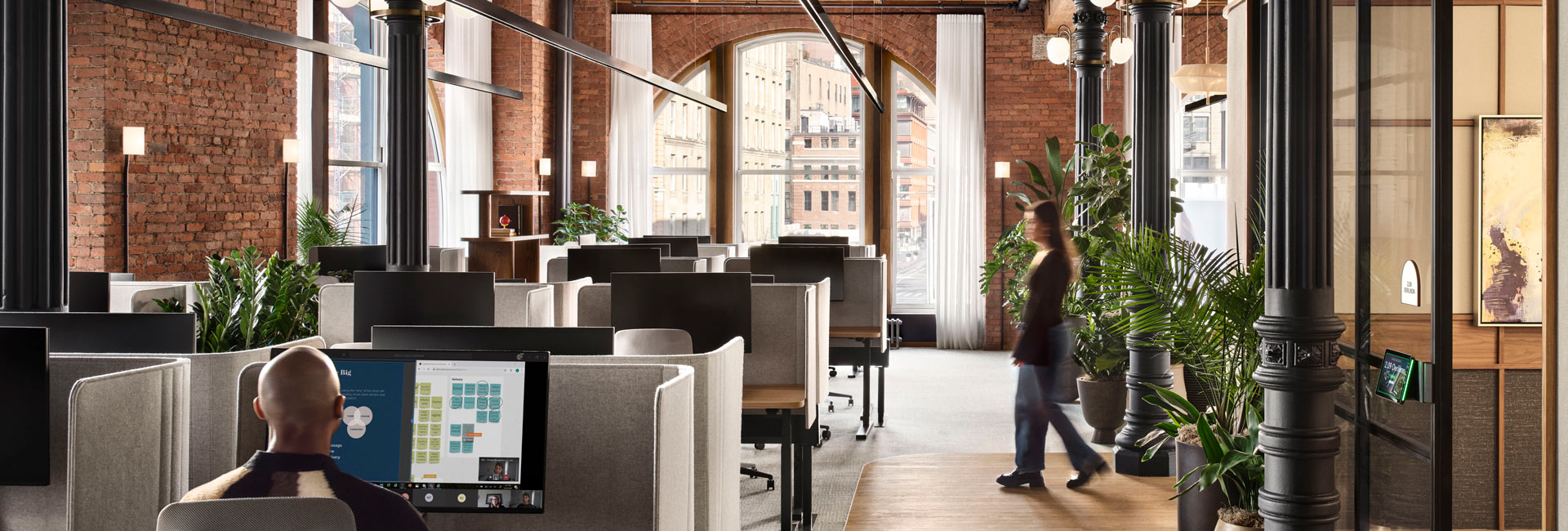 Plant-filled lounge and work area with sofas, tables and enclosed workstations beneath exposed wood ceilings and large arched windows.