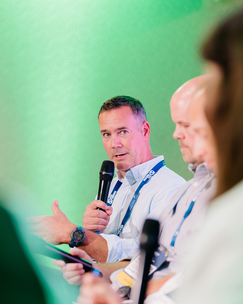 Andy Elliot speaking into a handheld microphone during a panel discussion, with other seated panelists in the foreground.