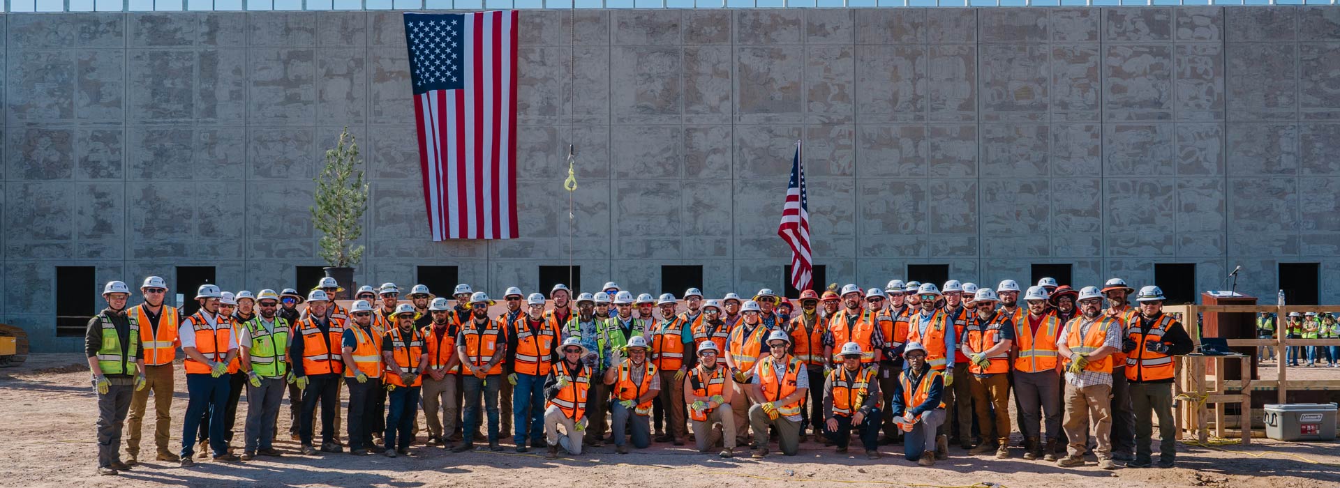 Large group of construction workers in hard hats and high-visibility vests posing in front of an unfinished concrete building with two American flags.